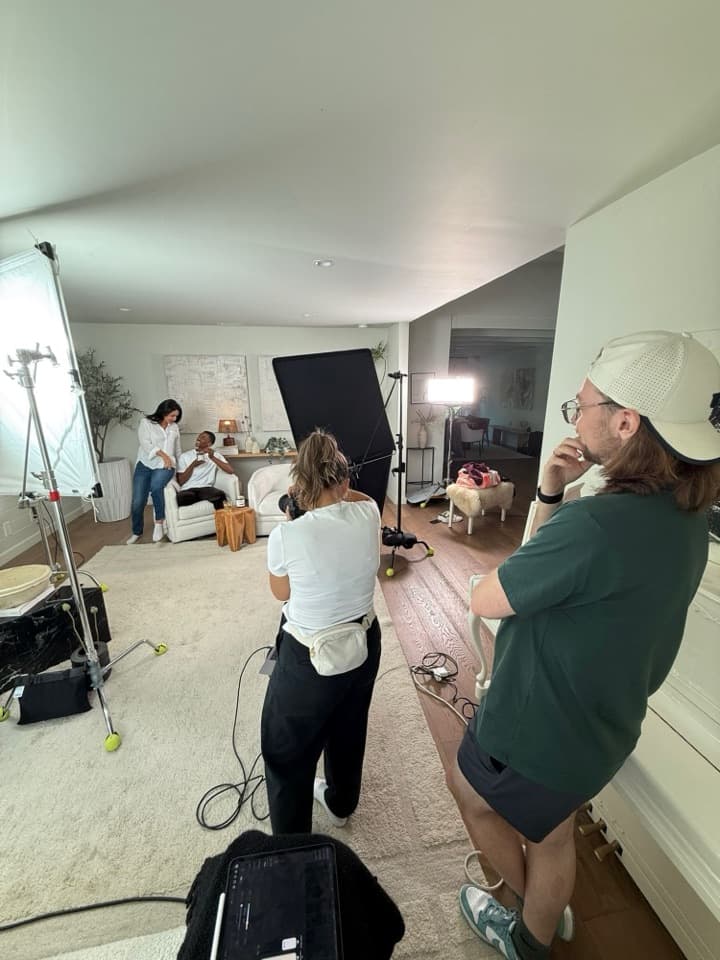 A living-room set lit through a large diffusion frame, with a photographer shooting two people on a sofa while a producer watches.