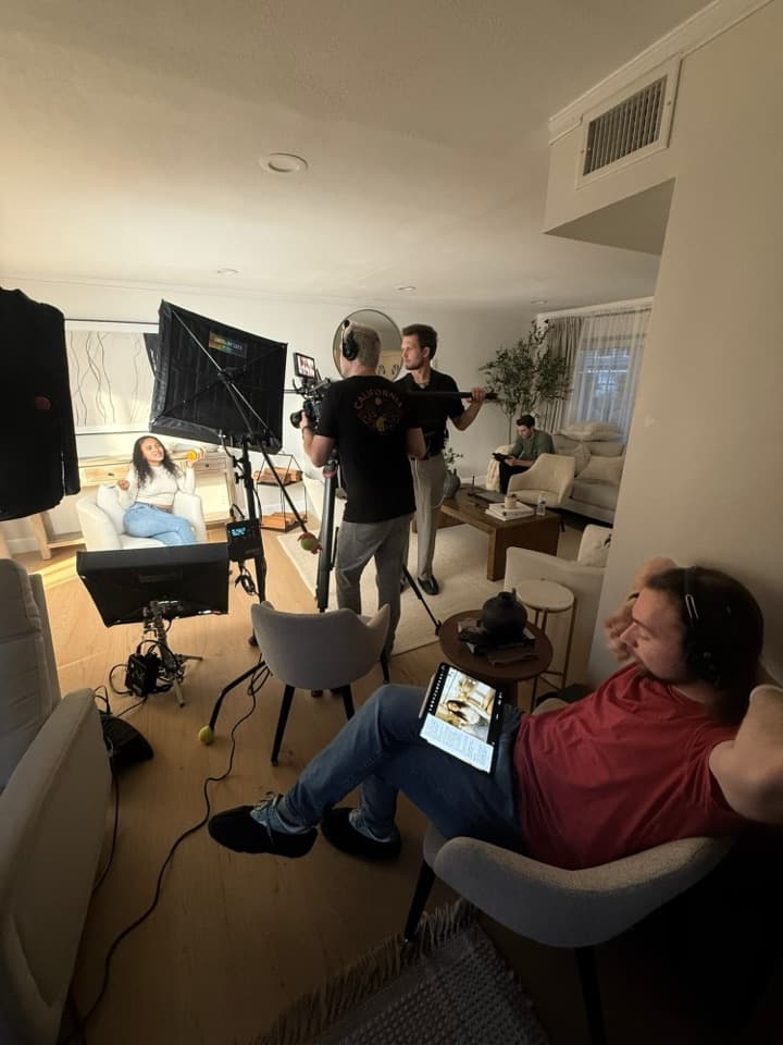 A living-room interview with softbox lighting, a camera operator and a boom operator, and a producer following the script on a tablet.